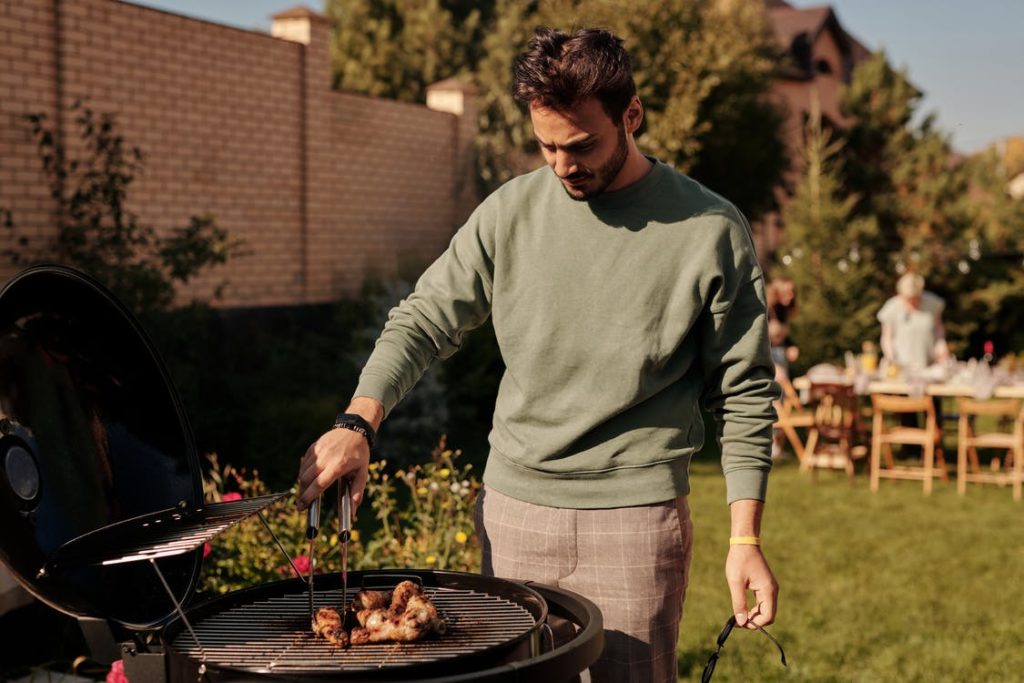 a man, who is white and has short brown hair and a beard, is wearing a green jumper and checked trousers in his garden. He is using prongs to turn over some chicken on a barbecue. Behind him is his wife, child, and mother, who are setting up a table outdoors.