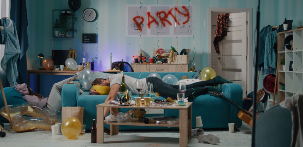 a male student is laying face first down on a sofa. Surrounding him are numerous empty glasses and food and rubbish across the room. Above, across a photo frame, the word "party" has been spray painted on in red paint.   